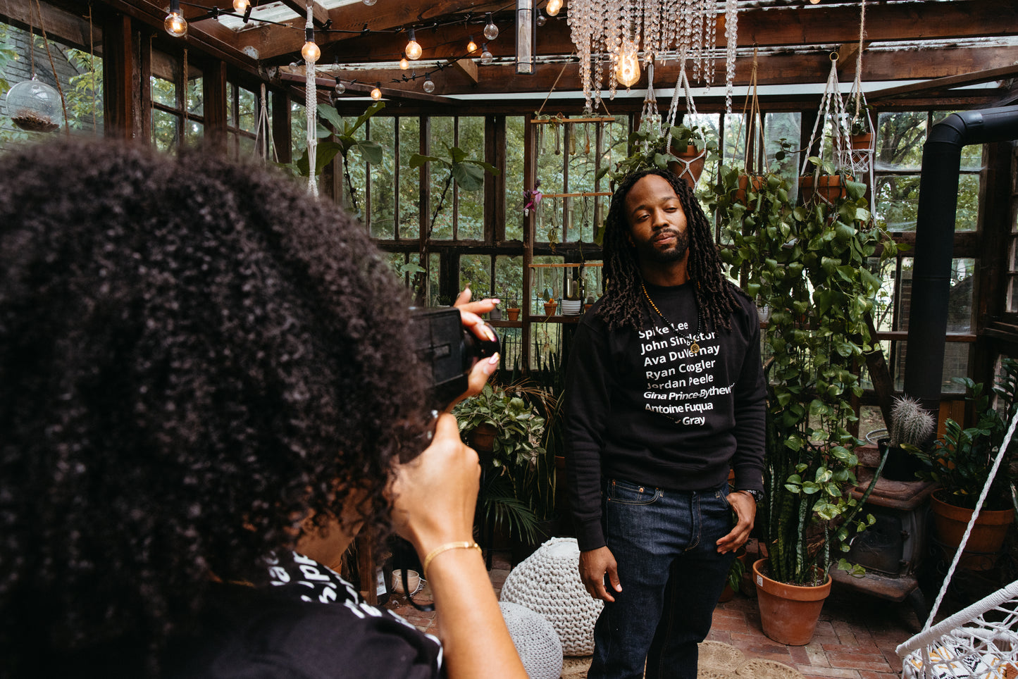 woman filming man wearing crew neck sweater with minimalist typography listing black filmmakers and cinema directors: Spike Lee, John Singleton, Ava DuVernay, Ryan Coogler, Jordan Peele, Gina Prince-Bythewood, Antoine Fuqua, F. Gary Gray